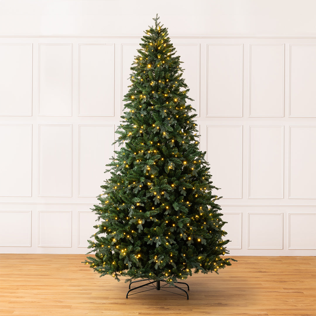 Christow Pre-Lit 10ft Fraser Fir Artificial Christmas Tree with warm white LED lights on a black metal stand on a light wood floor against a white panelled wall