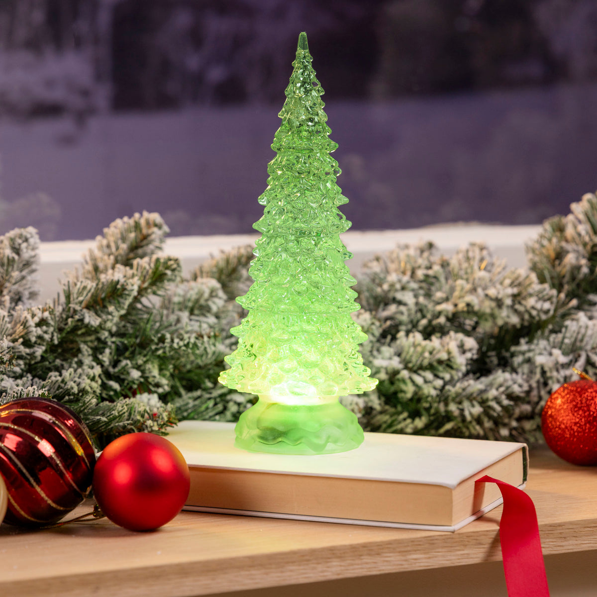 Christow Swirling Glitter LED Christmas Tree glowing green beside frosted greenery and Christmas baubles